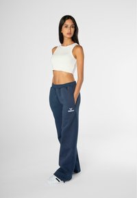 Top corto blanco de canalé combinado con pantalones de chándal azul marino con bolsillos laterales y bordado de logo. La modelo lleva zapatillas deportivas casuales.