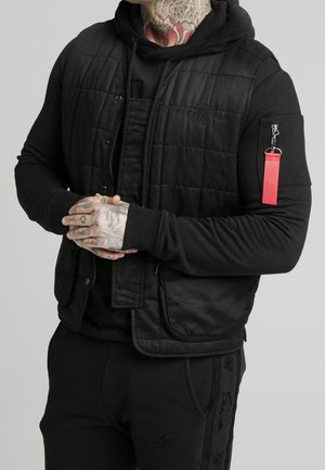 Mann mit tätowierten Händen und Nacken, trägt eine schwarze gesteppte Weste über einem schwarzen Hoodie mit rotem Reißverschlussband am Ärmel, die Hände gefaltet.