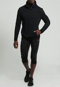 Felpa nera con cappuccio e colletto alto, abbinata a leggings sportivi neri a vita alta con righe laterali e sneakers nere.