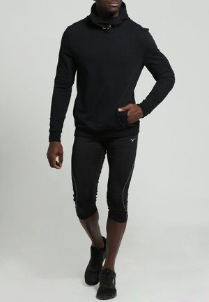 Sweat à capuche noir avec un col haut, associé à des leggings de sport noirs mi-longs avec des rayures sur les côtés, et des baskets noires.