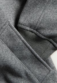 Grijze fleece stof met een gestructureerde afwerking; heeft een geribde manchet en een zijvak. Dichtbijopname van de naden en stikdetails.