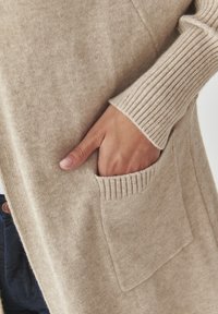 Cardigan beige en tissu doux, avec des poignets côtelés et une poche à l'avant. Texture notoire avec une finition lisse.