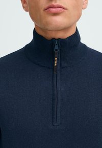 Pulover cu fermoar de culoare bleumarin, cu anevoiu și guler înalt, având un eticheta cu logo portocalie. Textura țesăturii este moale și ajustată.