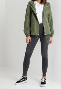 Donna con giacca a cappuccio verde oliva, t-shirt bianca, jeans skinny grigi scuri e sneakers nere e bianche, in piedi contro uno sfondo neutro.