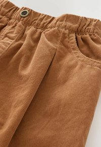 Pantaloni in velluto a coste marroni con vita elasticizzata, pieghe e una tasca laterale, con una texture di tessuto visibile su tutta la superficie.