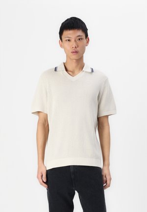 Kortermet, beige strikket polo skjorte med v-hals, ribbestrikket krage med marineblå striper. Brukt med mørke denimbukser.
