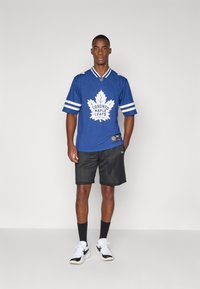 Blå kortermet jerseyskjorte med hvit Toronto Maple Leafs-logo og striper, kombinert med svarte mesh-shorts og hvite joggesko.