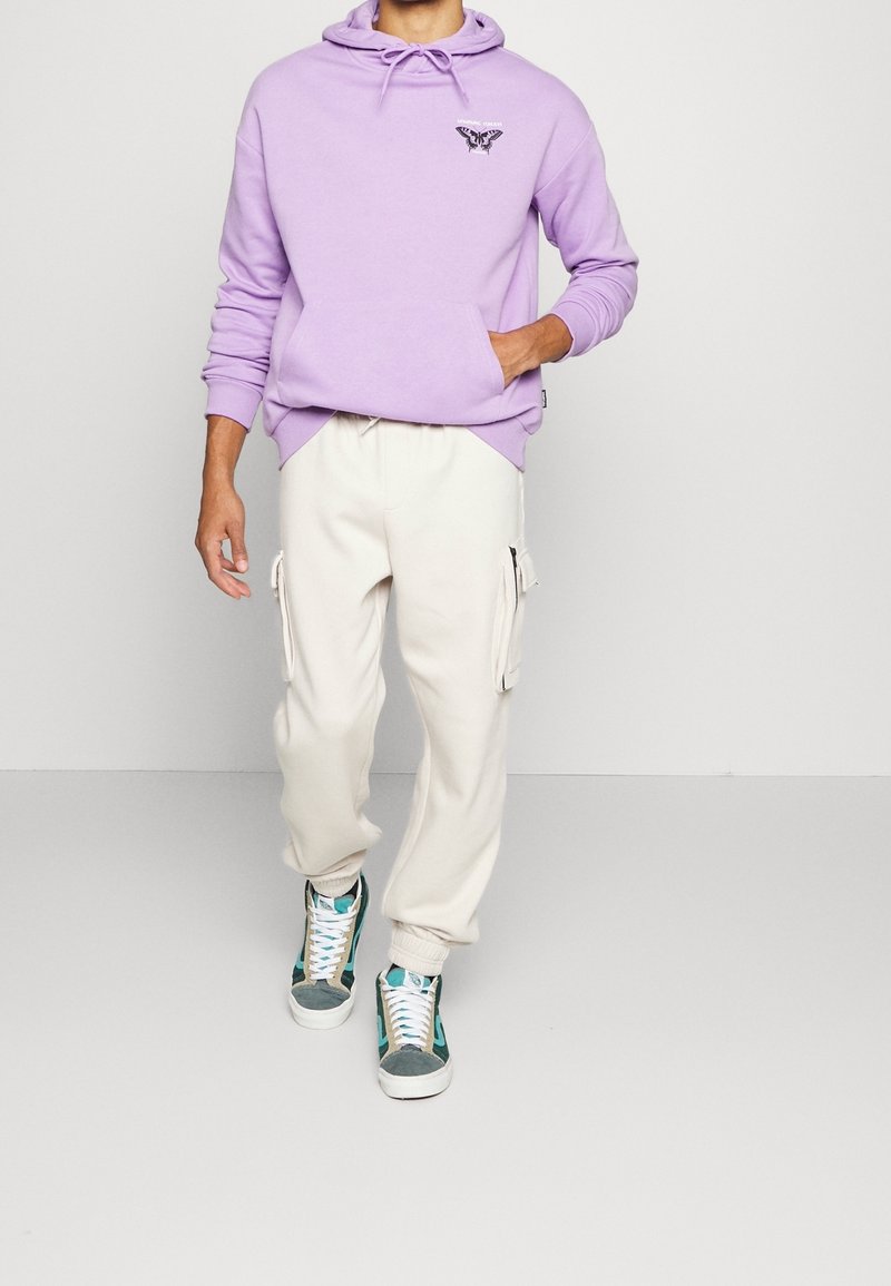 Lila hoodie med ett grafiskt fjärilstryck på bröstet, i kombination med beiga cargobyxor med sidofickor. Sportskor med turkosa accenter.