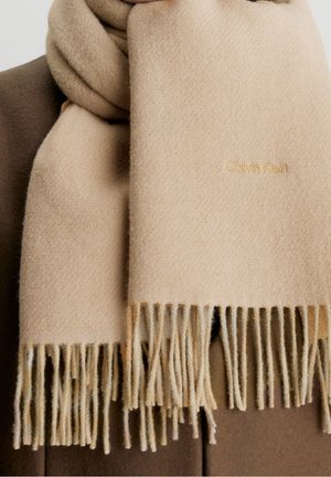 Écharpe en laine beige aux extrémités effilochées, enroulée autour d'une personne portant un manteau marron, avec une broderie discrète du logo Calvin Klein.