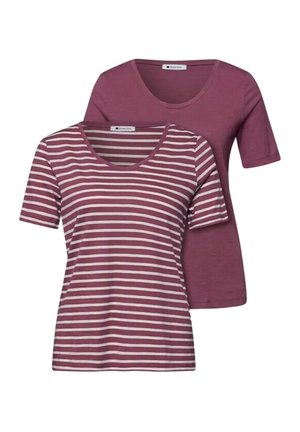 Dos mujeres están apoyadas contra una pared; una con una camisa malva a rayas y vaqueros azules sujetando un bolso marrón, la otra con una camisa malva y vaqueros grises recortados sujetando una chaqueta.