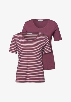 Deux femmes sont adossées à un mur ; l'une porte une chemise mauve rayée avec un jean bleu et tient un sac marron, l'autre porte une chemise mauve avec un jean gris court et tient une veste.