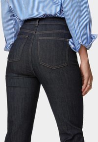 Los jeans de mezclilla oscura presentan una cintura de altura media, dos bolsillos traseros y costuras naranjas en contraste. Se combinan con una camisa a rayas azul y blanca.