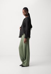 Blusa nera a maniche lunghe con vestibilità ampia, abbinata a pantaloni olive verdi a vita alta e gamba larga, indossati con décolleté neri a punta.