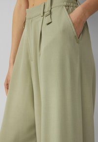 Šviesiai žali, plačios kojos kelnės iš tekstūruoto audinio, su priekinėmis raukšlėmis, šoninėmis kišenėmis ir reguliuojamu liemeniu su sagties detalėmis.