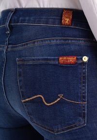 Dark blue denim jeans featuring a distinctive zigzag stitch in gold thread on the back pocket and a red brand label above.