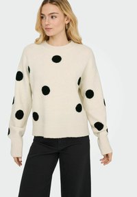 Pull crème à gros pois noirs, encolure ronde, manches longues et ourlet côtelé. Texture douce, coupe décontractée. Porté avec un pantalon noir.