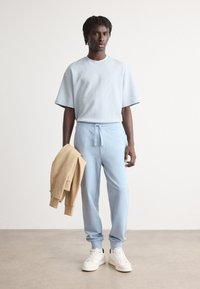 Lyseblå kortermet strukturert skjorte og matchende joggebukser, kombinert med hvite sneakers. En beige genser er lagt over den ene armen.