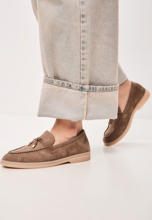 Brown suede loafers with a rounded toe, decorative tassels, and a soft beige sole, paired with light blue cuffed jeans.