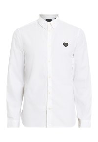 Camisa blanca de manga larga hecha de algodón, con un cuello abotonado y un logo de corazón negro bordado en el lado izquierdo del pecho.