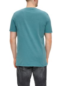 Teal kortlopende t-shirt, ronde halslijn, gladde textuur, met een kleine grafische print op de onderste linker achterkant, gedragen met donkere jeans.