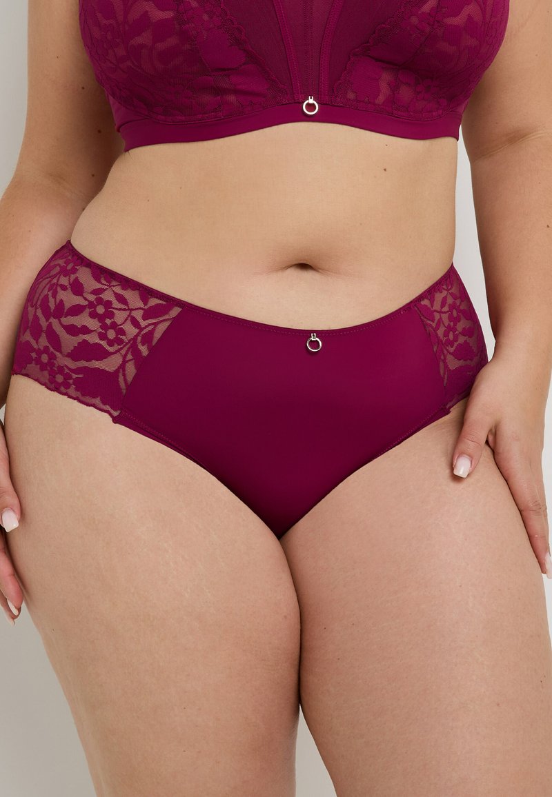 Femme taille plus portant un ensemble coordonné de soutiens-gorge en dentelle bourgogne et de culottes taille haute avec des panneaux en dentelle florale, se tenant avec les mains reposant sur les cuisses.