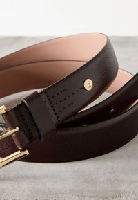 Tumepruun nahast vöö kuldse klambri ja väikese ümara kuldse logostudiga, rullitud kujul heledal hallil pinnal.