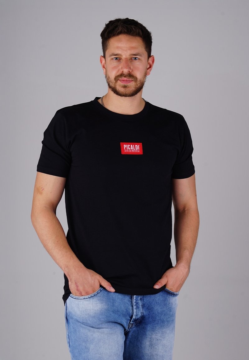 Mann mit Bart, der ein schwarzes T-Shirt mit rotem "PICALDI"-Logo und hellblauen Jeans trägt, steht mit den Händen in den Taschen vor einem grauen Hintergrund.