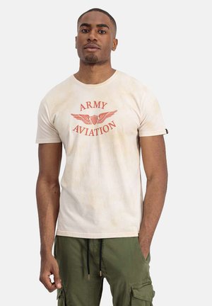 Junger Mann trägt ein hellbeiges T-Shirt mit der Aufschrift "Army Aviation" und einem geflügelten Emblem, olivgrüne Cargohose, steht mit einer Hand in der Tasche.
