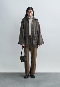 Bruine wollen mantel met franjes en grote zakken, gecombineerd met een lichte coltrui, bruine broek en een groene handtas. Zwarte schoenen maken de look compleet.