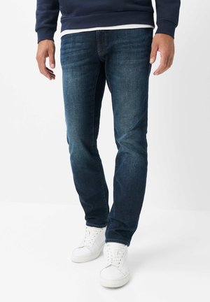 Mann trägt dunkelblaue Jeans, weiße Sneakers und ein marineblaues Sweatshirt, steht vor einem schlichten weißen Hintergrund.