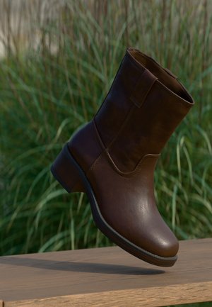 Brown leather ankle boot with low heel resting on wooden surface against green grass background.