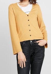 Gul ribbad långärmad kofta med v-hals, knappstängning och en kort design, matchad med svarta jeans.