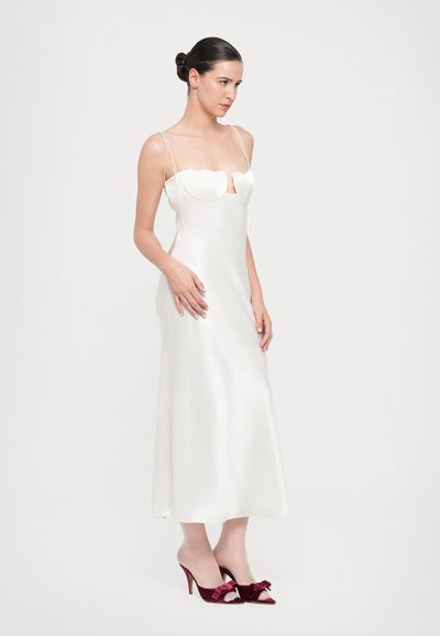 Vestido de satén blanco con escote en forma de corazón, tirantes finos y una silueta fluida. Combinado con tacones de terciopelo burdeos con lazos.