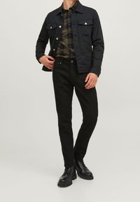 Chaqueta de mezclilla negra con botones plateados, combinada con una camisa de cuadros verde y negra. Unos pantalones ajustados negros y botas negras con cordones completan el conjunto.