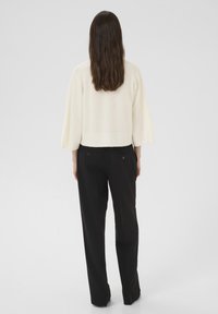 Hosszú barna hajú nő fehér, széles ujjú pulóverben és fekete, egyenes szárú nadrágban, egy sima háttér előtt áll.