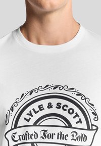 Camiseta de algodón blanca con un destacado estampado gráfico negro que incluye el texto "LYLE & SCOTT" y un diseño de borde ornamentado. Cuello redondo.