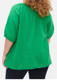 Persona con capelli ricci che indossa una blusa strutturata di un vivace verde brillante con maniche a tre quarti e pantaloni scuri, vista da dietro.