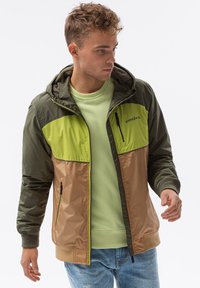 Färgglad jacka med olivgröna och tan paneler, zippered front, justerbar huva och elastiska ärmslut. Lätt, smidig textur.