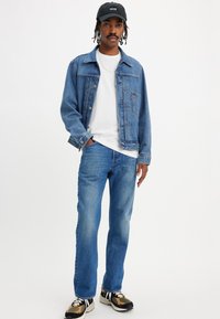 Veste en denim bleu moyen, t-shirt blanc à col rond en dessous, jeans bleu clair et baskets noires avec des accents beiges, habillé de manière décontractée.