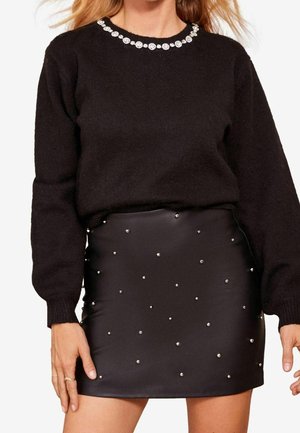 Kvinde iført en sort sweater med dekorative hvide perler på halsudskæringen og en sort miniskirt med spredte sølvnitter.