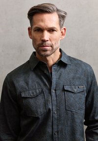 Ciemnoniebieska koszula w wzory wykonana z miękkiego materiału, z zapięciem na guziki z przodu, dwoma kieszeniami na piersiach i klasycznym kołnierzem.