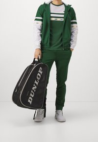 Zelený sportovní teplákový komplet s bílými akcenty, bunda na zip a odpovídající kalhoty. Drží černou tašku Dunlop se šedým logem a zipy.