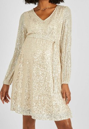 Frau trägt ein langärmeliges goldfarbenes Paillettenkleid mit V-Ausschnitt und Taillengürtel, steht vor einem schlichten Hintergrund.