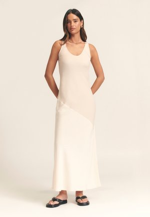 Sleek, cream-colored maxi dress with a V-neckline and thin straps, featuring a diagonal design. Paired with black flat sandals.