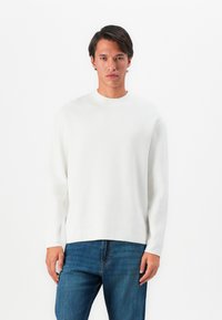 Hombre con cabello oscuro vestido con un suéter blanco de manga larga y jeans azules, de pie contra un fondo blanco.