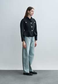 Kvinne stående iført svart knappet peplumjakke, lyseblå vide bukser og svarte loafers på grått gulv med lys grå bakgrunn.