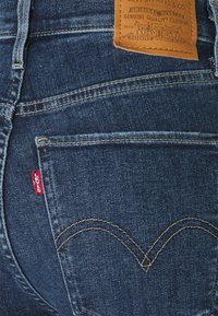 Jeans de mezclilla azul oscuro con costuras visibles y una etiqueta de cuero que muestra los detalles de la marca. Etiqueta roja en el bolsillo trasero izquierdo. Diseño clásico.