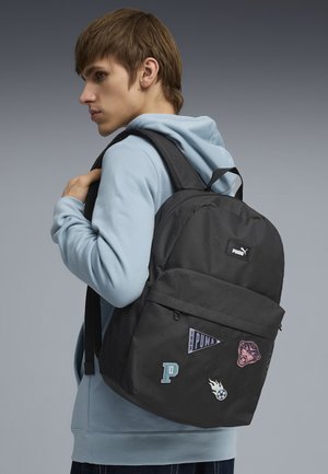 Jeune personne portant un sweat à capuche bleu clair, avec un sac à dos noir Puma orné de patches, regardant sur le côté sur un fond gris.