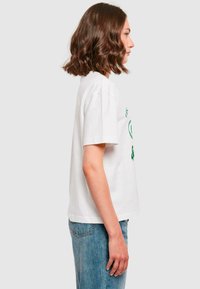 T-shirt en coton blanc à manches courtes, avec un texte imprimé vert sur la poitrine. Coupe décontractée, associé à un jean en denim bleu.