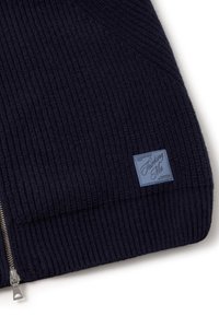 Marineblauwe geribbelde gebreide trui met een zilveren ritssluiting en een blauw merklipje. Heeft een verticale textuur en een aansluitend ontwerp.